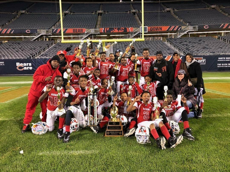 Youth football team in red uniforms and coaches pose with trophies on a football field.