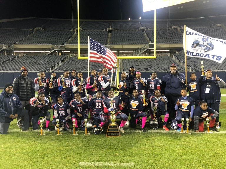 Youth football team poses with trophies and US flag on field after championship game.
