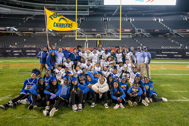 Youth football team and cheerleaders pose with trophies on a football field. A "FISC Chargers" flag is held aloft.