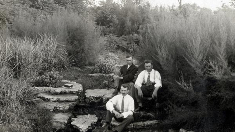 Black and white photo of three men sitting on rocks in a garden.