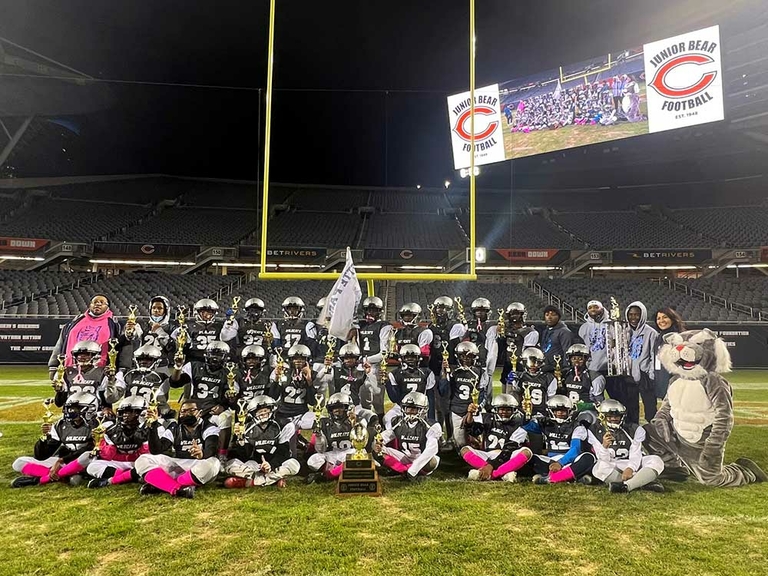 Junior football team poses with trophies on a field. A large screen displays team photo.