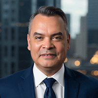 Headshot of a smiling man in a blue suit and white shirt against a city view background.