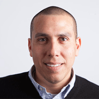 Headshot of a smiling man in a shirt and sweater against a white background.