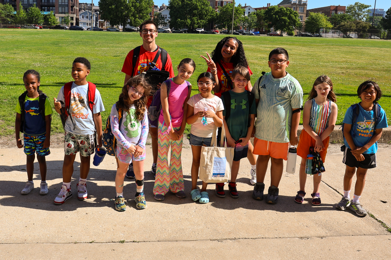 Group of children and a counselor pose for a photo on a sidewalk next to a grassy field.