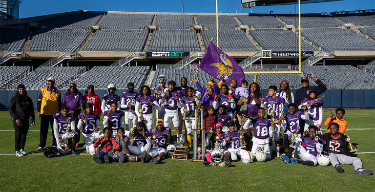Youth football team photo, on a field, holding trophies and a Vikings flag.2024 Junior Bear Football Champs in the end zone at Soldier Field.