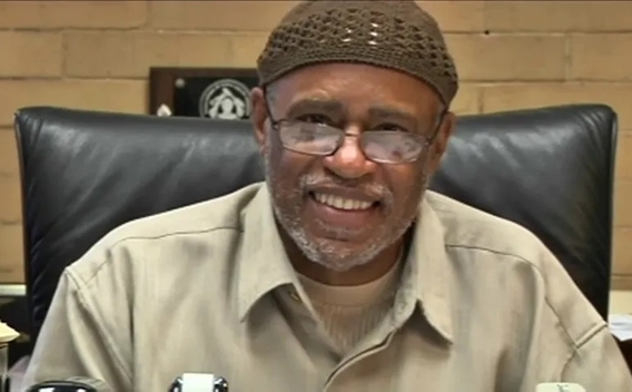 Smiling photo of Dr. Conrad Worrill, wearing glasses and a knit hat, sitting at a desk smiling.