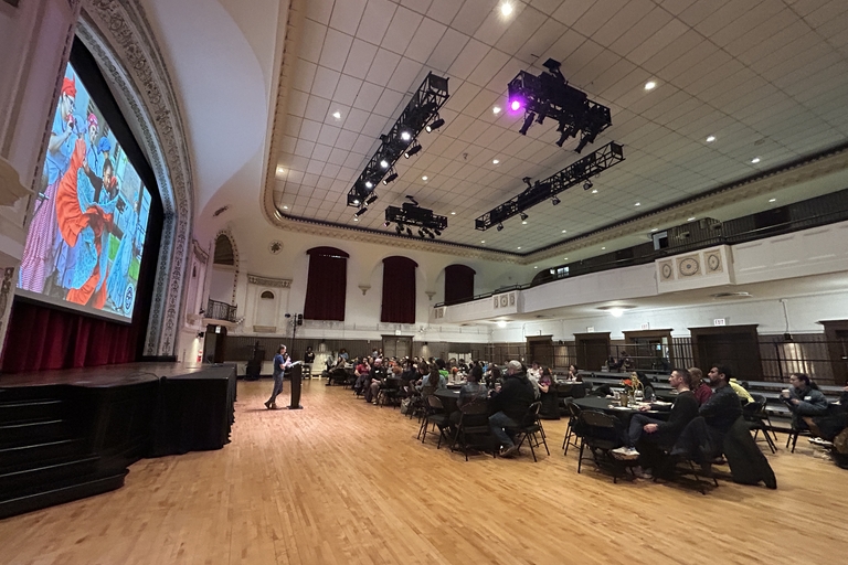 A speaker presents to an audience seated at round tables in a large auditorium. A projected image of people in colorful clothing is visible on a screen behind the stage.