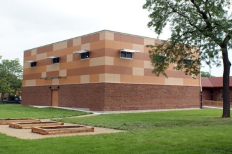 Modern two-story school building with brick lower level and tan and orange upper level. Raised garden beds in foreground.