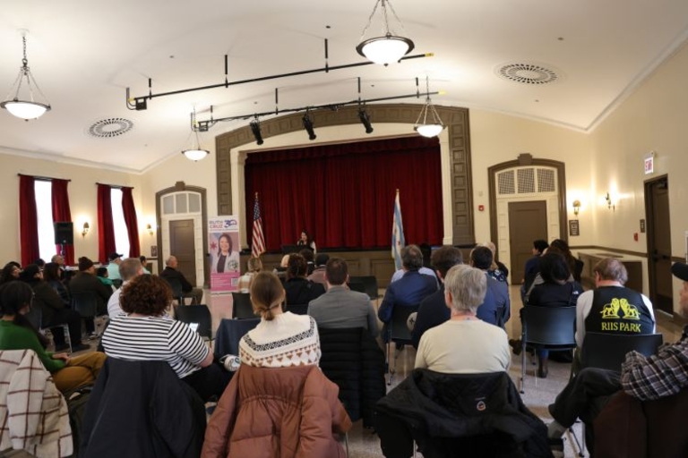 Audience sits in chairs facing a stage with a speaker and two flags.