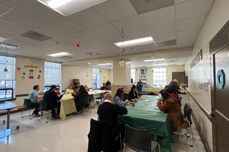 People sit at tables eating at a birthday party in a community room.