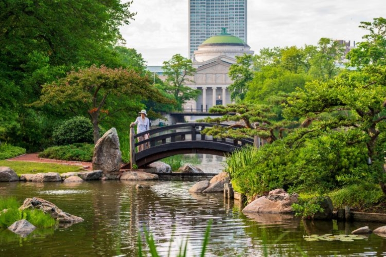 Woman on arched footbridge in peaceful Japanese garden. Building and skyscraper in background.