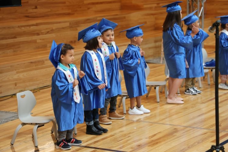 Preschoolers in blue graduation gowns stand on a wooden gymnasium floor.