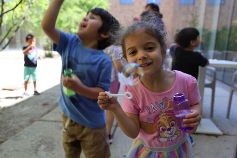 Girl blowing bubbles, smiling at the camera.  A boy in the background reaches for a floating bubble.