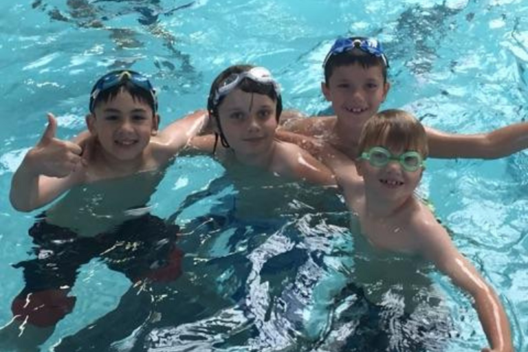 Four children wearing goggles smile in a bright blue swimming pool. One gives a thumbs up.