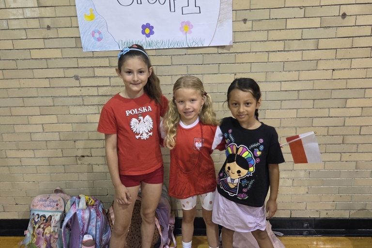 Three girls wearing red and white, representing Poland, stand in a gymnasium by a brick wall. One holds a small Polish flag.