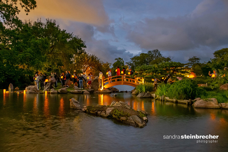 People with lanterns gather on bridge and shores of pond in a Japanese garden at twilight.