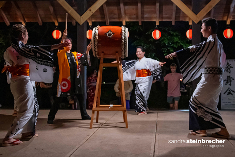 Four Japanese drummers in yukata, two playing, two dancing, at a nighttime festival.