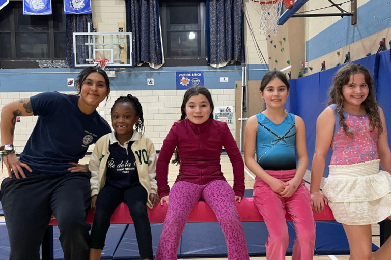 Adult woman and four smiling girls in a gym, sitting on a pink beam.