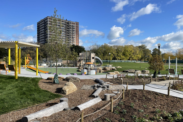 Photograph of a sunny public playground with play equipment, grassy areas, and a city apartment building in the background.