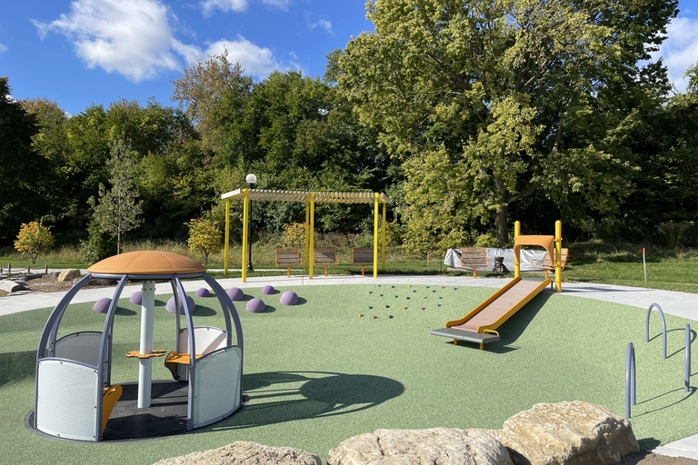 A brightly lit, modern playground with a carousel, small slide, and a wooden pergola.