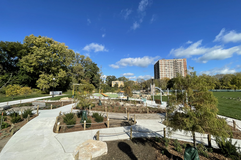 Photograph of a sunny park with concrete pathways, landscaping, and a playground under a blue sky.