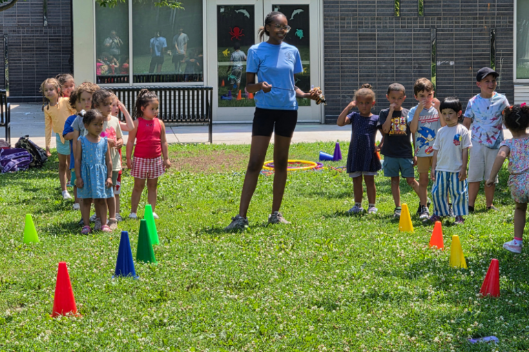 A young woman stands on a grassy lawn with a group of young children and colorful cones.