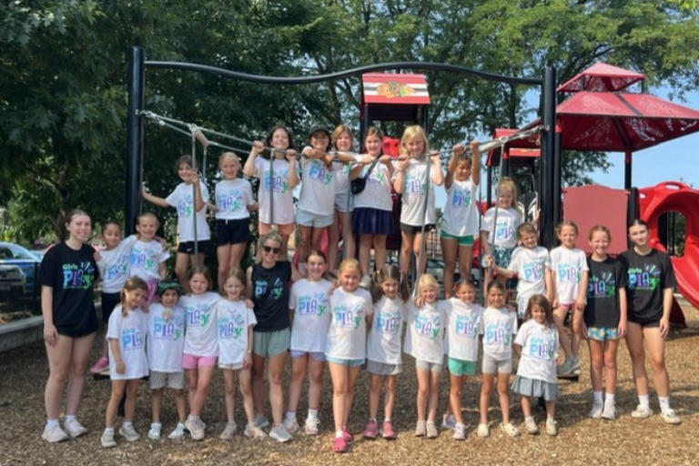 A large group of girls wearing "Girls Play" t-shirts posed together at a sunny playground.