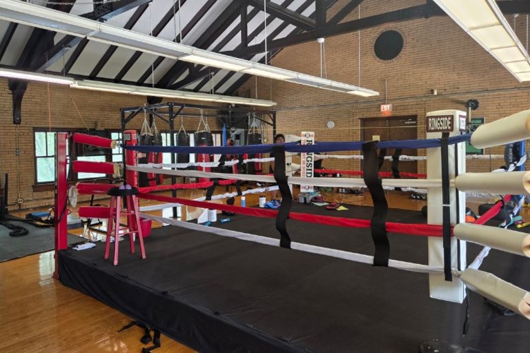 Boxing ring with padded ropes in a gym with brick walls and exposed ceiling beams.