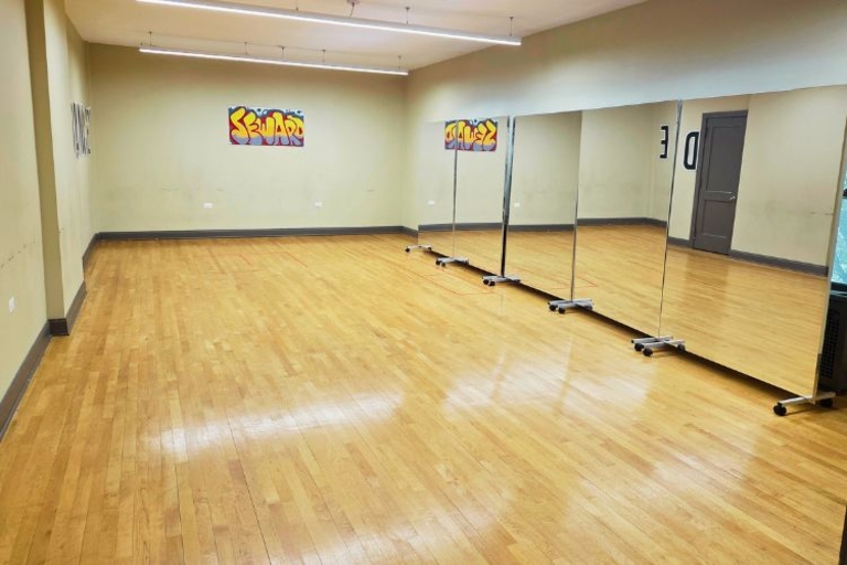 Photograph of an empty dance studio with wooden floors, fluorescent lights, and rolling mirrors.