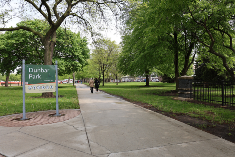 Two people walk along a path in a lush, green Dunbar Park on a sunny day.