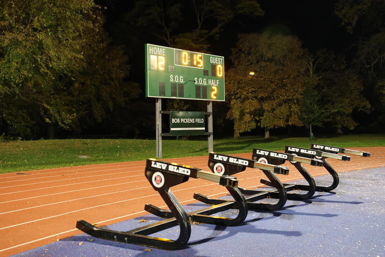 A line of Lev Sleds on a running track at night with a digital scoreboard in the background.