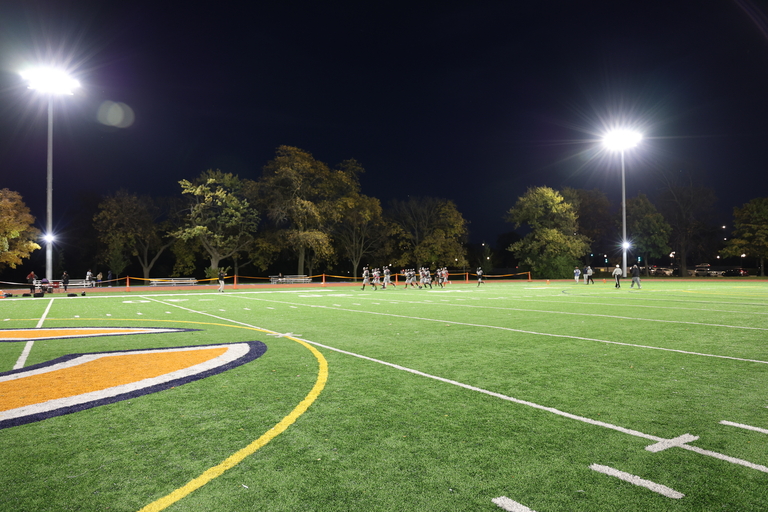 Photograph of a football field at night illuminated by stadium lights with players in the distance.