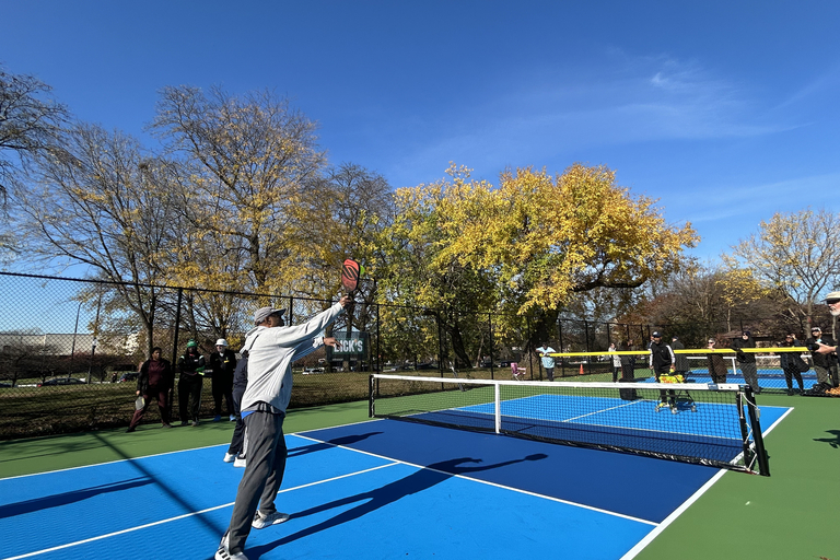 A man hits a pickleball on a vibrant blue court outdoors under a clear sky with autumn trees.