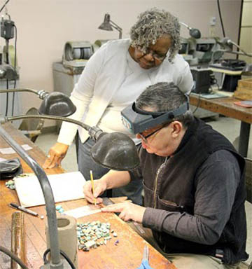Lapidary artist sketching a design with turquoise pieces nearby, instructor observes.