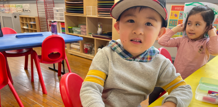 Boy wearing a backwards baseball cap sits at a table in a classroom as a girl in the background puts a drawing over her head.