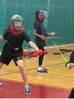 Two fencers wearing protective gear practice in a gym.