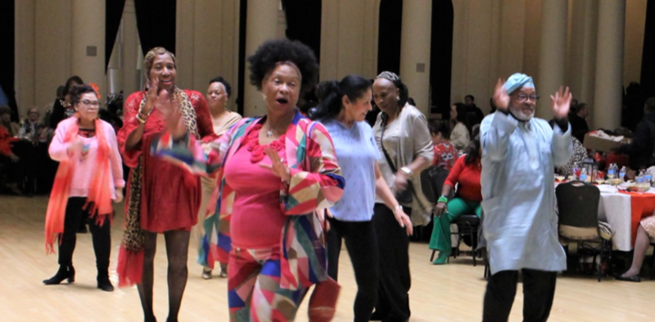 A group of people dance and wave their hands in a ballroom. Tables with red and white tablecloths are visible in the background.