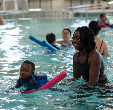 Tiny Tot Swim - Level 1.png Smiling woman and toddler with flotation device in pool during swim class.