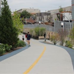 People stroll and jog on a paved urban path lined with greenery.