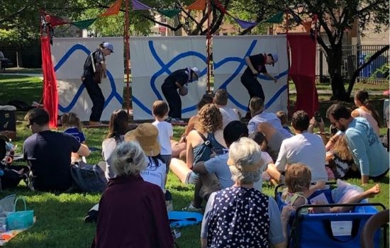 Outdoor children's theater performance with audience seated on grass. Three performers paint on large canvases.
