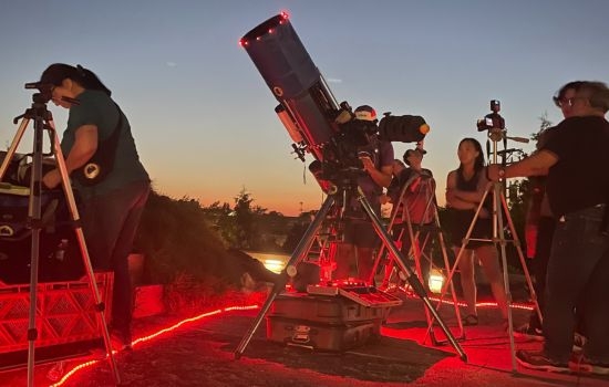 People use telescopes and cameras at dusk, illuminated by red lights.