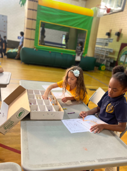 Two girls coloring at a table with crayons and an inflatable bounce house in the background.