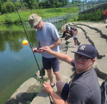 Boy smiles, holding a fishing rod with a small fish caught, as man takes a photo.