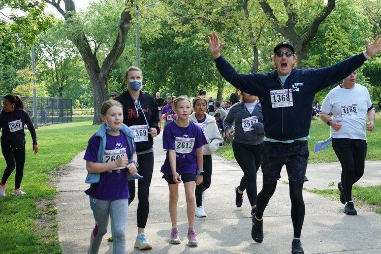 Runners in purple shirts jog on a paved path through a park. One runner raises his arms in excitement.