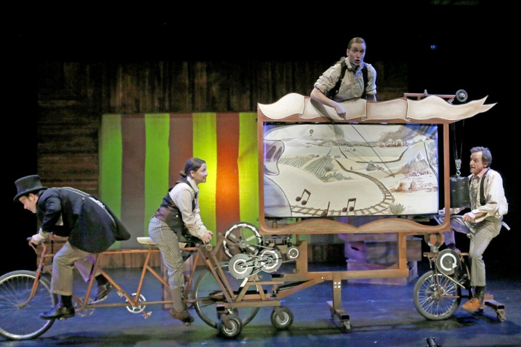 Four vaudeville performers operate a bicycle-powered rolling illustrated display of a train moving through a landscape. Music notes float above the train tracks.
