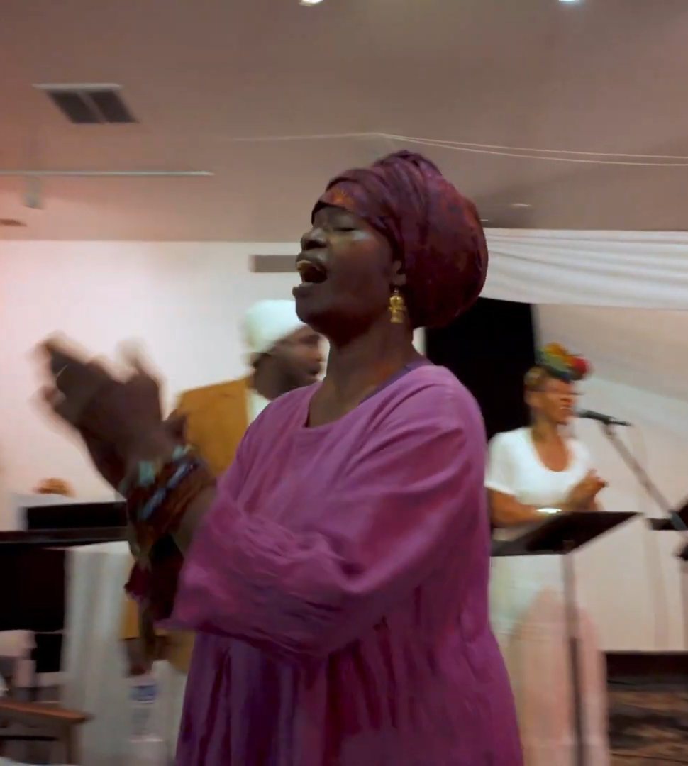 A woman in a magenta dress and headwrap sings and claps.