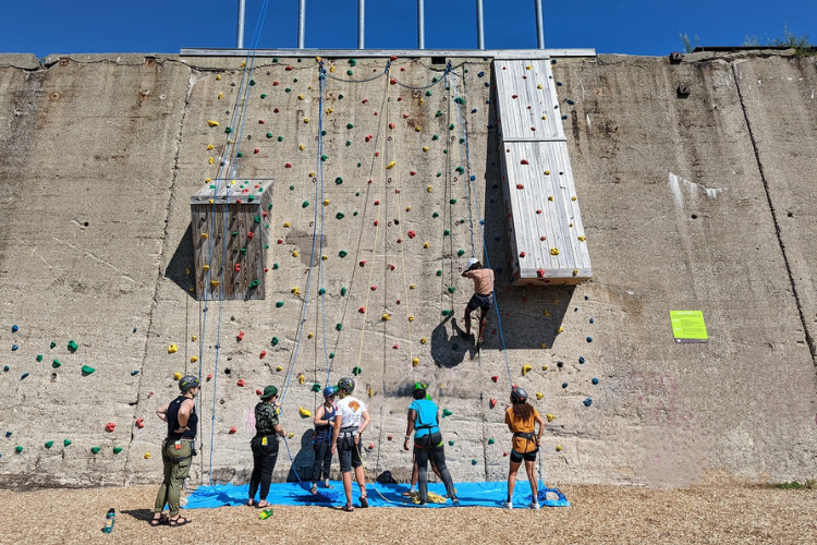 Group of people rock climbing outdoors on a concrete wall with colorful handholds. A blue safety mat lies on the ground below.