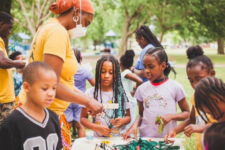 Children and an adult work on a craft project at an outdoor table.