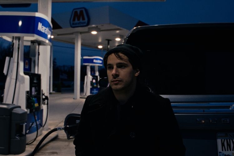 A man leans against the back of his car at gas station at night.