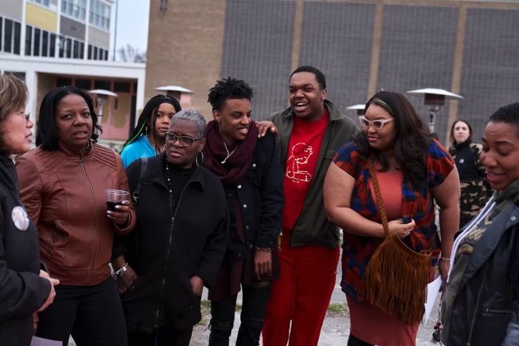 A group of community organizers stand together outside a school building, talking and smiling.